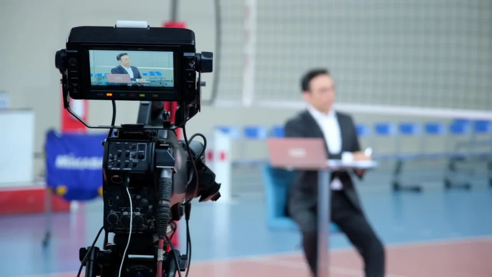 Caméra de plateau TV en tournage : sur l’écran de contrôle, un présentateur assis à une table avec un ordinateur portable, dans un hall de sport aménagé en studio